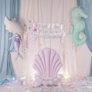 May include: A light pink seashell shaped chair with white balloons around the base. A mermaid balloon with a purple tail and a green seahorse balloon are in the background. A banner with the words "Happy Birthday" is hanging above the chair.