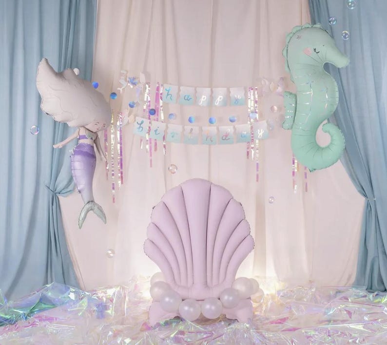 May include: A light pink seashell shaped chair with white balloons around the base. A mermaid balloon with a purple tail and a green seahorse balloon are in the background. A banner with the words "Happy Birthday" is hanging above the chair.