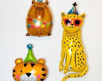 May include: Three animal-shaped foil balloons: a brown capybara with a party hat and banner, a yellow cheetah with sunglasses, and an orange tiger with a green party hat. All balloons are against a white background.