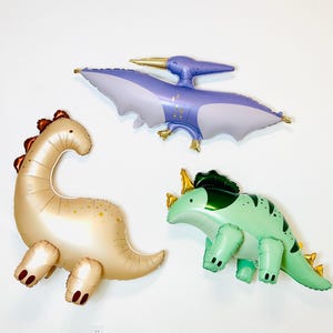 Puede incluir: Tres globos de dinosaurio inflables. Un pterod&aacute;ctilo morado, un brontosaurio marr&oacute;n y un triceratops verde.
