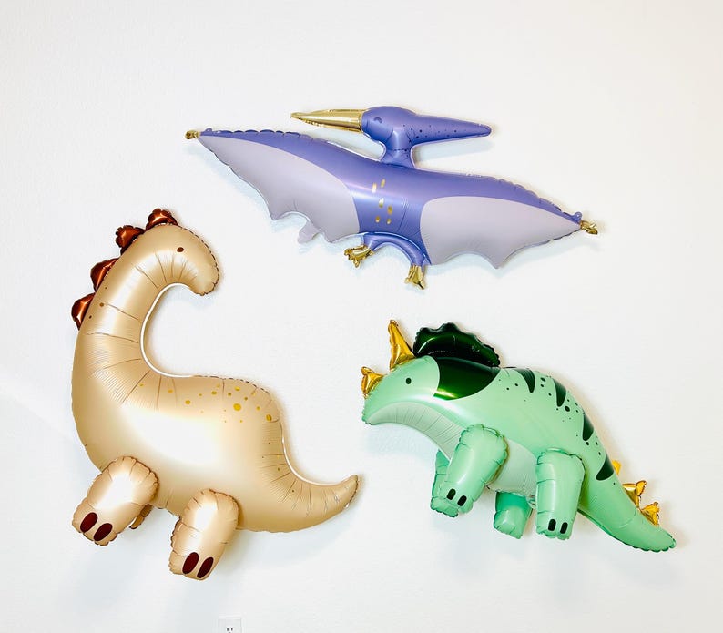 Puede incluir: Tres globos de dinosaurio inflables. Un pterod&aacute;ctilo morado, un brontosaurio marr&oacute;n y un triceratops verde.