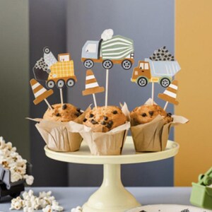 May include: A set of four cupcakes decorated with construction vehicle themed toppers. The cupcakes are on a white cake stand. There is a small toy dump truck in the foreground.