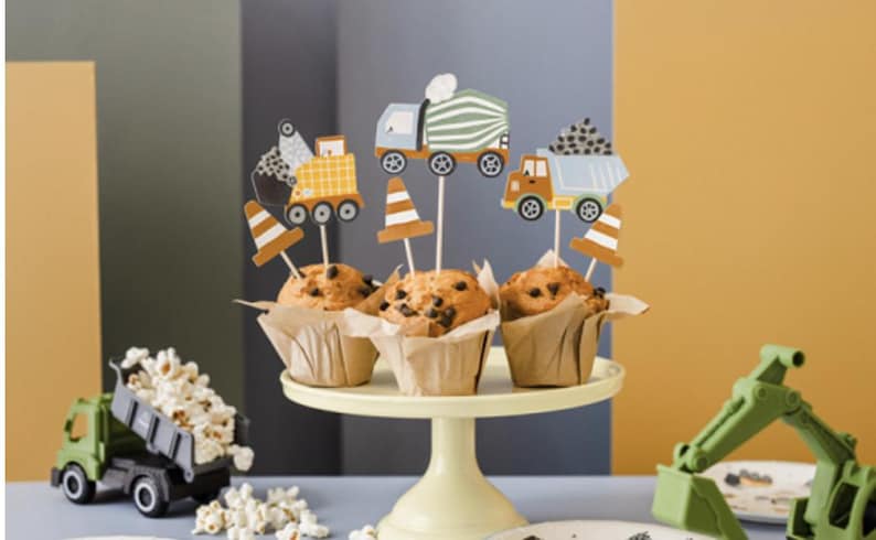 May include: A set of four cupcakes decorated with construction vehicle themed toppers. The cupcakes are on a white cake stand. There is a small toy dump truck in the foreground.