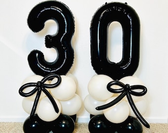 Puede incluir: Dos globos negros con el número 30 sobre grupos de globos en blanco y negro. Cada grupo está coronado con un lazo negro. Los globos están colocados contra una pared blanca y una alfombra beige.