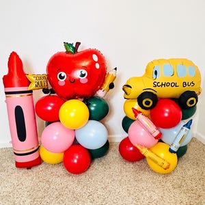 May include: Two balloon arrangements featuring foil balloons shaped like a pink crayon with a sign reading SCHOOL RULES, a red apple with a face, and a yellow school bus. These are accented with round red, yellow, pink, blue, and green balloons.