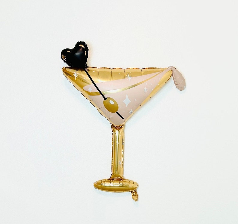 K&ouml;nnte beinhalten: Goldfarbener Folienballon in Form eines Martini-Glases mit rosafarbenem Inneren und goldenen Glitzer. Der Ballon hat einen schwarzen herzf&ouml;rmigen Anh&auml;nger oben.
