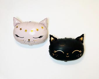 May include: Two cat-shaped foil balloons, one pink with gold stars and one black with gold eyes and a heart nose.