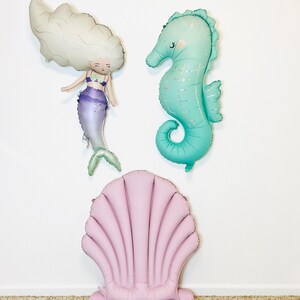 May include: A set of three inflatable decorations featuring a mermaid, a seahorse, and a seashell chair. The mermaid is white with purple and green accents. The seahorse is turquoise with a glittery finish. The seashell chair is pink.