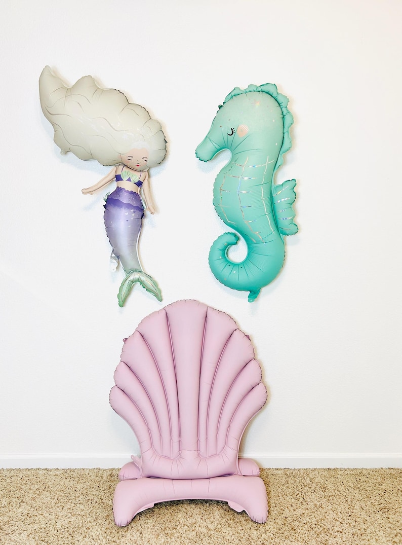 May include: A set of three inflatable decorations featuring a mermaid, a seahorse, and a seashell chair. The mermaid is white with purple and green accents. The seahorse is turquoise with a glittery finish. The seashell chair is pink.