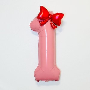 May include: A pink number one foil balloon with a shiny red bow attached at the top. The balloon is inflated and has a smooth surface. The bow is made of a shiny red material. The balloon is likely for a first birthday or anniversary.