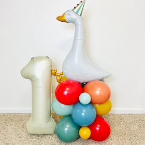 May include: A festive balloon arrangement for a first birthday celebration. It features a large silver number one balloon, a white goose balloon wearing a party hat, and a cluster of colorful balloons in red, blue, orange, and yellow.