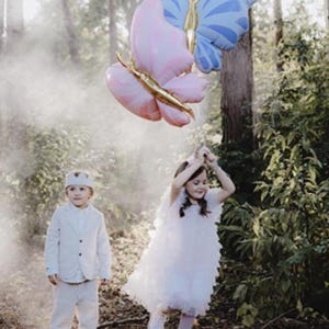 May include: Two children, one wearing a white suit and a crown, and the other wearing a white dress, stand in a forest with two large butterfly balloons. The balloons are pink and blue with gold accents.