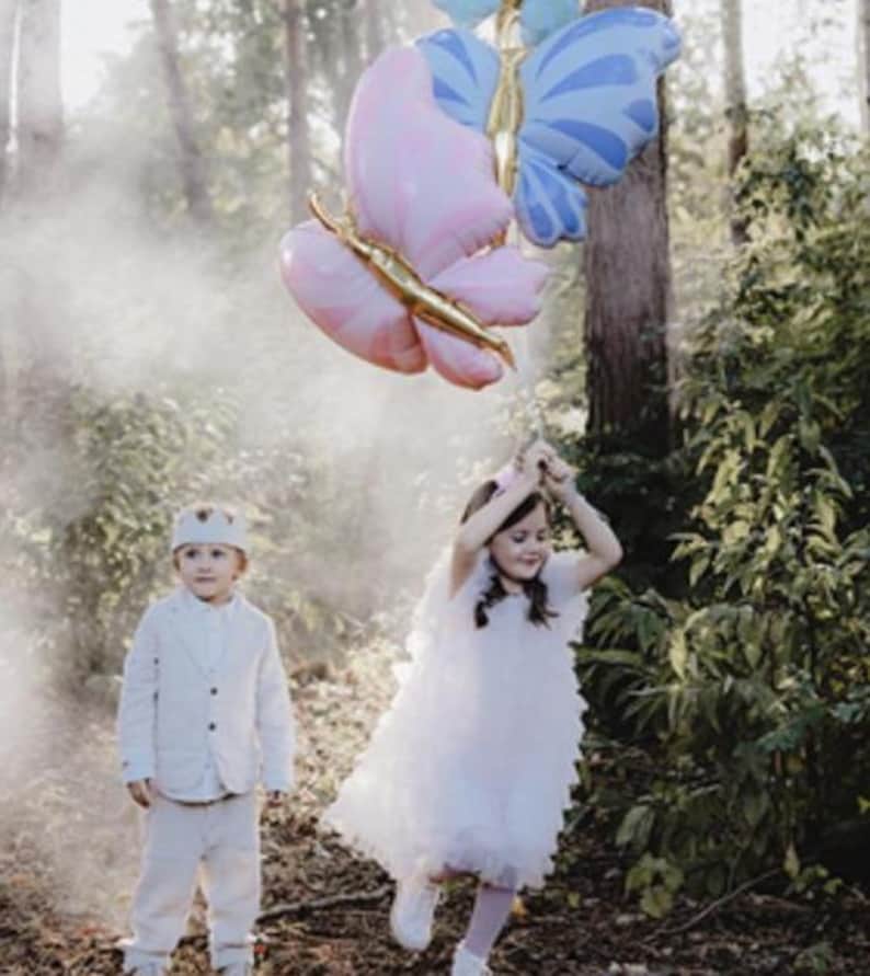 May include: Two children, one wearing a white suit and a crown, and the other wearing a white dress, stand in a forest with two large butterfly balloons. The balloons are pink and blue with gold accents.