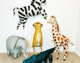 Può includere: Una collezione di cinque palloncini in alluminio a forma di animale, tra cui una zebra bianca e nera, un elefante grigio, un suricato giallo, una giraffa marrone e crema e un coccodrillo verde scuro, disposti come decorazioni gonfiabili.