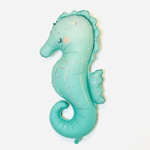 May include: A teal blue seahorse shaped balloon with a silver glitter design. The balloon has a cute face with closed eyes and a small pink nose.