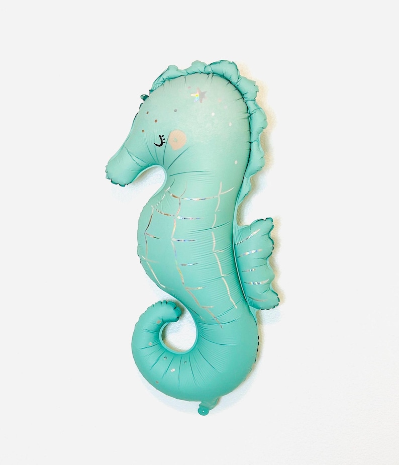 May include: A teal blue seahorse shaped balloon with a silver glitter design. The balloon has a cute face with closed eyes and a small pink nose.