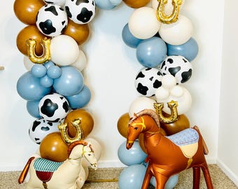 May include: A balloon arch featuring blue, brown, white, and cow-print balloons with gold horseshoe-shaped foil balloons. Two inflatable horse figures stand in front, one cream-colored with a green striped saddle and one brown with a blue saddle.