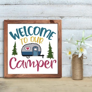 May include: A framed sign with the text "Welcome to our Camper" in a whimsical font. The sign features a cartoon camper with a heart on the side, two pine trees, and a pink and yellow color scheme.