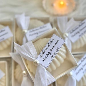 Könnte beinhalten: Weiße Muschelschalen-Seifen in transparenten Boxen, gebunden mit weißem Band und Perlenakzent. Jede Box hat ein Etikett mit dem Text "Catherine Mathew, First Holy Communion, 04.25.2026". Die Seifen sind auf einer weißen Oberfläche angeordnet.