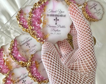 May include: White and gold round favor with pink glitter and a cross. The favor says "Thank You for celebrating with us!" and "Breanna Padilla Baptism 04.27.2025".