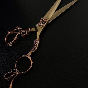 May include: Ornate copper-colored scissors with gold blades. The handles and finger holes are embellished with red gemstones and decorative patterns. A small frog-like figure adorns the pivot point. The scissors rest on a black surface.