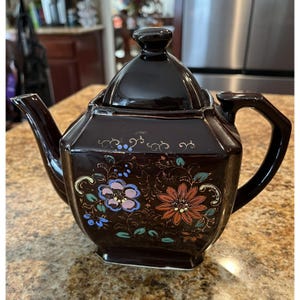 May include: A dark brown ceramic teapot with a matching lid. The teapot features floral designs in shades of blue, pink, orange, and gold. The teapot has a square shape with a handle and spout.
