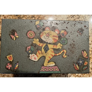May include: Vintage metal box with a cartoon tiger juggling colorful shapes and blocks. The box is painted in shades of green and features illustrations of flowers and butterflies. The tiger is yellow with orange stripes.