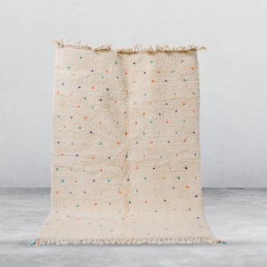 Puede incluir: Alfombra color crema con un patrón de puntos en varios colores. La alfombra tiene una superficie texturizada y bordes con flecos. Los puntos son en tonos de azul, naranja y verde. La alfombra se muestra sobre un fondo neutro.