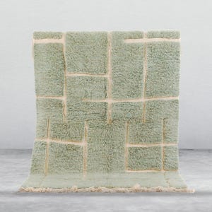 May include: A rectangular, light green rug with a geometric design. The rug features a series of textured, rectangular blocks in varying sizes, separated by off-white lines. The rug has a soft, plush appearance and a fringed edge. This rug is suitable for home decor.