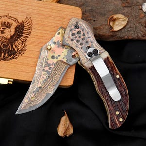 Peut inclure: Un couteau de poche pliant avec une lame en acier damas à motifs présentant des accents de cuivre et de laiton. Le manche est fabriqué en bois de cerf brun texturé avec un clip de poche en métal argenté. La lame possède un bouton de pouce.