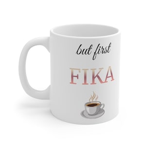 Może przedstawiać: Biały ceramiczny kubek do kawy z napisem "but first FIKA" w czarnych i różowych literach. Pod tekstem znajduje się mała ilustracja filiżanki do kawy i spodka. Kubek ma zakrzywiony uchwyt.