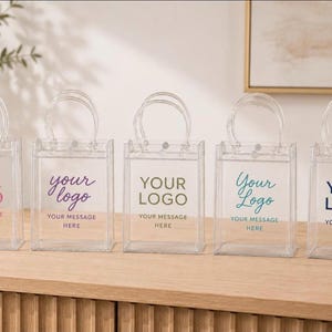 Peut inclure: Cinq sacs fourre-tout rectangulaires en plastique transparent avec des poignées transparentes. Chaque sac présente le texte imprimé YOUR LOGO et YOUR MESSAGE HERE en différentes couleurs : rose, violet, vert olive, bleu sarcelle et bleu marine.
