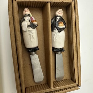May include: A set of two metal spreaders with ceramic handles shaped like waiters in black jackets and white aprons. One waiter holds a bottle, the other holds a small food item. The utensils are presented in a rectangular brown cardboard box.