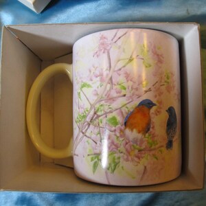 May include: A white ceramic mug with a handle, featuring a floral design with two bluebirds. The birds are perched on branches with pink blossoms.