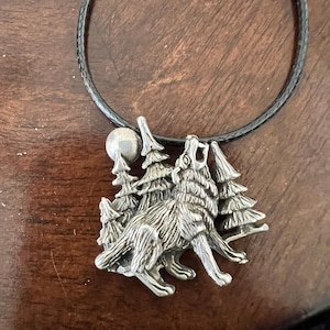 May include: A silver pendant necklace featuring a wolf howling at the moon with trees in the background. The necklace is on a black cord.