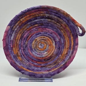 May include: A handmade, circular trivet with a spiral design. The trivet is made of fabric in shades of purple, orange, and pink, with a loop handle. The trivet is approximately 7 inches in diameter, and is suitable for hot dishes.