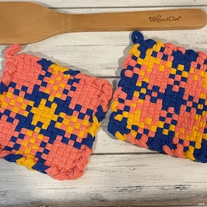 May include: Two woven pot holders in a pink, yellow, and blue geometric pattern. The pot holders are on a white wooden surface.