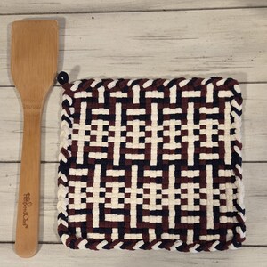 May include: A woven square hot pad with a geometric pattern in white, navy, and burgundy. A wooden spatula with the text "Pampered Chef" is next to the hot pad. The hot pad has a loop for hanging.