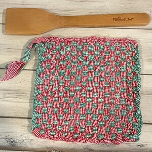 May include: A red and green checkered woven pot holder with a wooden spatula with the text "Pampered Chef" on it.