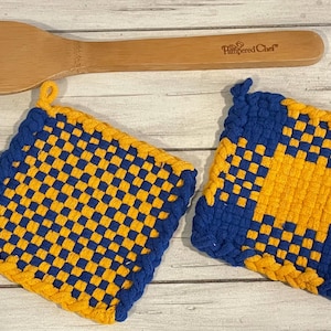 May include: Two blue and gold woven pot holders with a checkered pattern. The pot holders are made of cotton yarn and have a fringed edge.