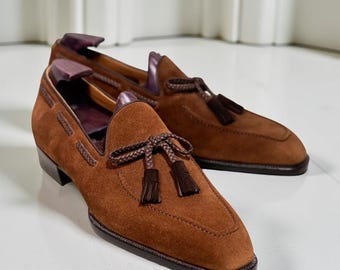 Op de afbeelding: Een paar bruine suède loafers met kwastjes. De schoenen hebben een vierkante neus en een donkerbruine zool. De binnenkant is donkerder bruin. De schoenen worden op een wit oppervlak gepresenteerd.