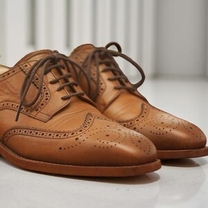 K&ouml;nnte beinhalten: Ein Paar hellbraune Leder-Brogue-Schuhe mit braunen Schn&uuml;rsenkeln. Die Schuhe haben dekorative Perforationen und stehen auf einer hellen Oberfl&auml;che. Klassischer Stil.