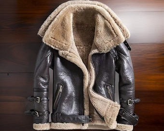 May include: A dark brown leather jacket with a shearling lining and collar. The jacket has a zip closure, two front pockets, and adjustable straps at the waist and cuffs. The jacket is displayed against a wooden background.