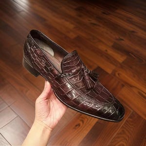 Peut inclure: Mocassin en cuir marron avec une texture peau de crocodile. La chaussure présente un détail de gland et un bout carré. La chaussure est tenue dans la main d'une personne.