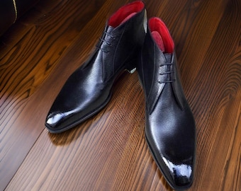 May include: A pair of black leather ankle boots with a glossy finish. The boots have a red interior and are laced up the front. The shoes are displayed on a wooden surface.