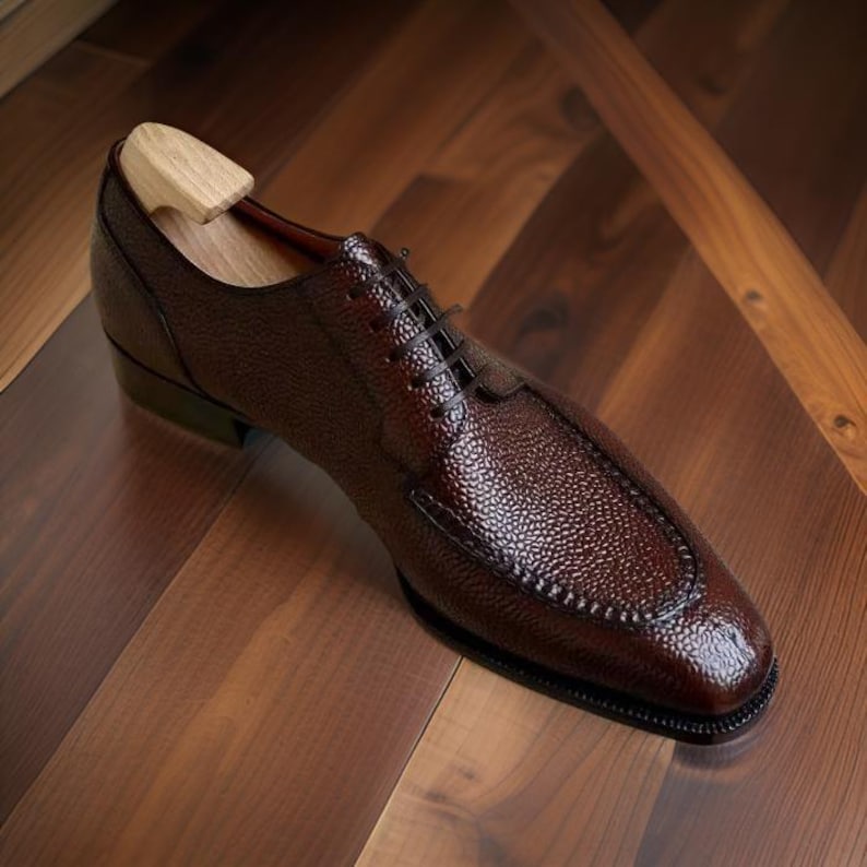 May include: A brown leather dress shoe with a textured surface and black laces. The shoe has a wooden shoe tree inside. The shoe is on a wooden surface.