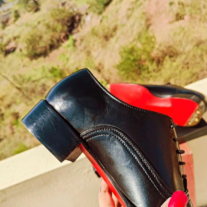 May include: Black leather dress shoes with red soles. The shoes have a low heel and a classic lace-up design. The soles are a vibrant red, contrasting with the black leather.