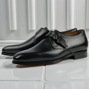 Könnte beinhalten: Ein Paar schwarze Lederschuhe mit Schnallenverschluss. Die Schuhe haben ein schlankes, poliertes Aussehen und einen niedrigen Absatz. Die Schuhe werden auf einer weißen Fliesenoberfläche präsentiert.