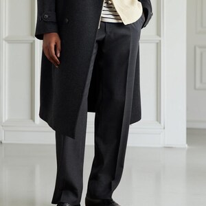 May include: A dark gray overcoat with a double-breasted design. The coat is worn over a cream-colored sweater and a striped shirt. The outfit includes dark gray trousers and black leather dress shoes.