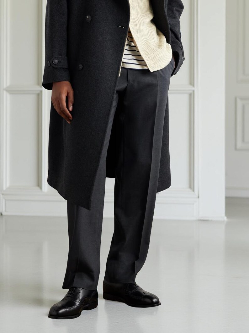 May include: A dark gray overcoat with a double-breasted design. The coat is worn over a cream-colored sweater and a striped shirt. The outfit includes dark gray trousers and black leather dress shoes.